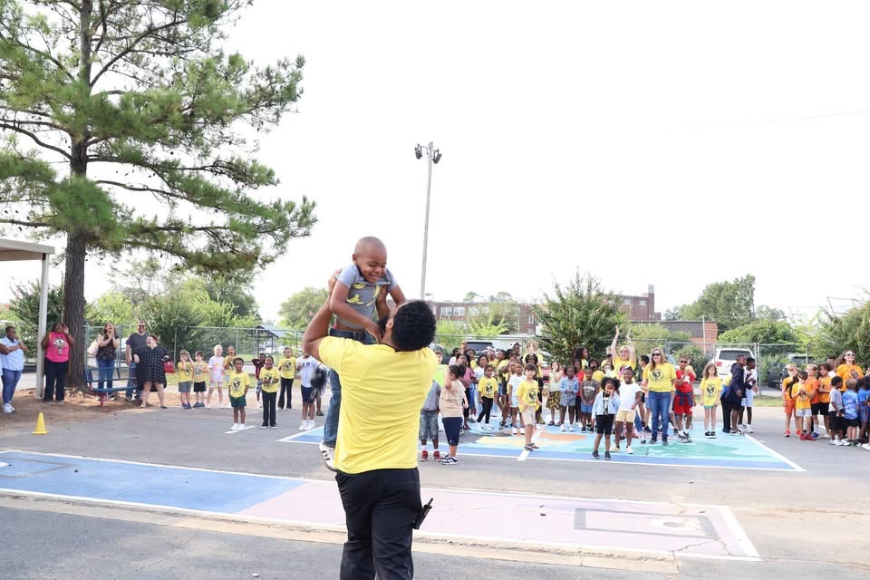 Gibbs students in yellow Laskavost house shirts cheering on the playground as an adult lifts a child on his shoulders.